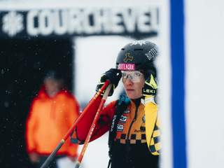 Ein Skirennläufer in Rennbekleidung und Helm stellt seine Skibrille ein, im Hintergrund ist ein unscharfes Courchevel-Schild zu sehen.