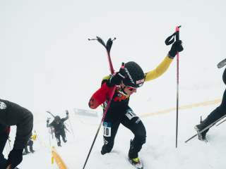 ein Athlet ist zu Fuß am Berg unterwegs, mit den Skiern auf dem Rücken. Im Hintergrund sieht man andere Athleten im Aufstieg und viel Nebel.