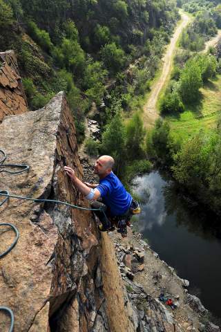 Ein Mann klettert gesichert an einer steilen Felswand, umgeben von einer grünen Landschaft mit einem Fluss und einem Wanderweg.