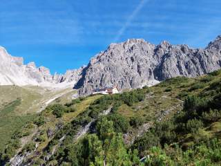 Berghütte auf einer grünen Alm vor steilen, felsigen Bergen unter blauem Himmel.