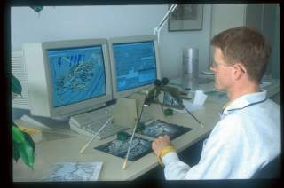 Ein Mann sitzt an einem Schreibtisch mit zwei Computerbildschirmen, auf denen geographische Karten zu sehen sind, und betrachtet mit einem Stereomikroskop großformatige Luftbilder oder Karten.
