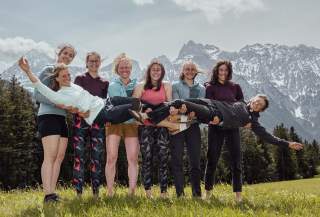 Sieben fröhliche Frauen stehen im Freien vor einer Berglandschaft und tragen gemeinsam eine achte Frau waagerecht in ihren Armen. Alle tragen sportliche Kleidung und lachen in die Kamera.
