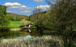 Kleines Haus am Ufer eines Teichs, umgeben von grünen Wiesen, Bäumen und einem bewölkten blauen Himmel.
