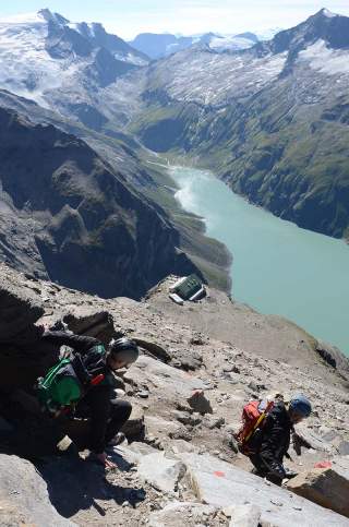 Zwei Bergsteiger mit Helmen und Rucksäcken klettern auf steinigem Pfad in hochalpiner Landschaft mit Blick auf einen langen, türkisfarbenen Bergsee und schneebedeckte Gipfel im Hintergrund.