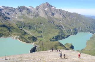 Wanderer steigen auf einem steinigen Bergpfad mit Blick auf einen Stausee und eine imposante Gebirgskette im Hintergrund. Das Wetter ist klar und sonnig.