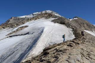 Zwei Wanderer mit Rucksäcken steigen auf einem schmalen, felsigen Grat in einem schneebedeckten Hochgebirge bei klarem Himmel auf.