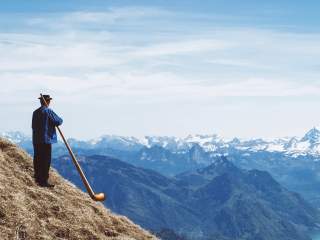 Mann in traditioneller Kleidung steht auf einer Bergwiese und spielt Alphorn, im Hintergrund eine Bergkette mit schneebedeckten Gipfeln unter blauem Himmel.