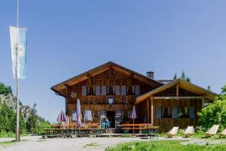 Holzhaus im alpinen Stil mit Außenbestuhlung, Sonnenschirmen und Liegestühlen vor blauem Himmel, umgeben von Bäumen und Bergen.