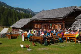 Menschen sitzen an Holzbänken und Tischen vor einer Berghütte in einer sonnigen Berglandschaft, während Hühner über die Wiese laufen. Im Hintergrund sind bewaldete Berge und ein blauer Himmel zu sehen.