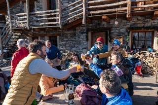 Eine Gruppe Menschen sitzt draußen an einem Holztisch vor einer rustikalen Steinlodges und stößt mit Biergläsern und Flaschen an, bei sonnigem Wetter. Im Hintergrund sind Holzstapel und eine Holztreppe zu sehen.