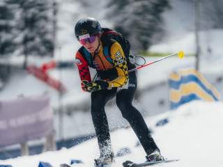 Eine Skimo Athletin in der Abfahrt auf Ski. Sie ist in einer gehockten Position, die Stöcke unter den Armen. Der Hintergrund ist verschwommen, man sieht Schnee und Zäune.