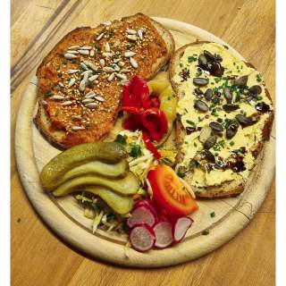 Holzbrett mit zwei Brotscheiben, eine bestrichen mit rotem Aufstrich und Sonnenblumenkernen, die andere mit hellgelbem Aufstrich, Kürbiskernen und Kräutern, dazu Gurkenscheiben, Radieschenscheiben, Tomatenstück, Paprikastreifen und Krautsalat.