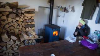 Eine Person sitzt in einem Raum neben einem brennenden Holzofen, umgeben von gestapeltem Feuerholz und verschiedenen Gegenständen auf einem Holztisch.