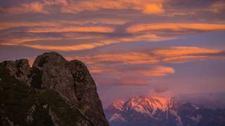 Berglandschaft im Abendlicht mit einem Felsen im Vordergrund und schneebedeckten Bergen im Hintergrund, unter einem dramatisch orange-violetten Himmel.
