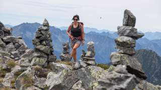 Frau mit Rucksack und Sonnenbrille wandert zwischen mehreren gestapelten Steinmännchen in einer bergigen Landschaft.