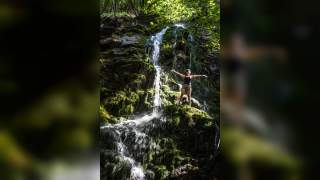 Frau steht mit ausgebreiteten Armen auf moosbedecktem Felsen vor einem kleinen Wasserfall in einem von Sonnenlicht durchfluteten Wald.