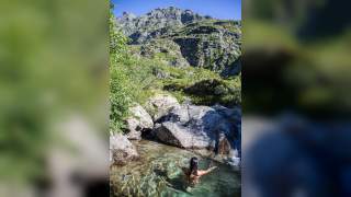 Person mit nassen Haaren badet in klarem Bergwasser, umgeben von Felsen und grüner Vegetation unter blauem Himmel.