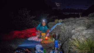 Frau liegt nachts in einem roten Schlafsack auf einer Felsfläche, trägt eine Stirnlampe und ist von Camping-Equipment und einer brennenden Kerze umgeben.