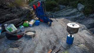 Outdoor-Kochstelle mit Campingkocher, Topf, Besteck, Lebensmitteln und Rucksack auf einem Felsen in einer naturbelassenen Umgebung.