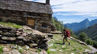 Wanderin mit rotem Rucksack nähert sich einer alten, aus Stein gebauten Hütte in einem bergigen, bewaldeten Gebiet unter blauem Himmel.