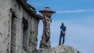 Ein Mann steht auf einem Felsen und blickt auf eine hölzerne Christusfigur mit Überdachung, daneben ist die Wand eines alten Steinhauses zu sehen.
