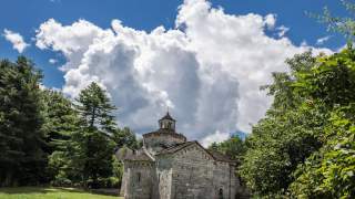 Alte steinerne Kapelle umgeben von grünen Bäumen unter einem blauen Himmel mit großen weißen Wolken.