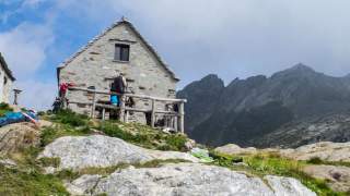 Steinernes Berghaus mit Personen, die auf Wiesen und Felsen inmitten von Bergen und bewölktem Himmel ruhen und entspannen.