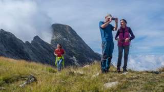 Drei Wanderer stehen auf einer grasbewachsenen Bergwiese mit felsigem Berg im Hintergrund, wobei ein Mann ein Foto macht und zwei Frauen ihn begleiten.