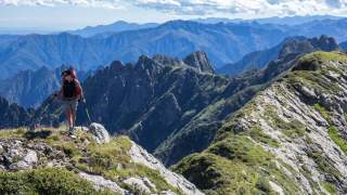 Eine Wanderin mit Rucksack und Wanderstöcken erklimmt einen felsigen, bewachsenen Berggrat, im Hintergrund sind weitere Bergketten unter blauem Himmel zu sehen.