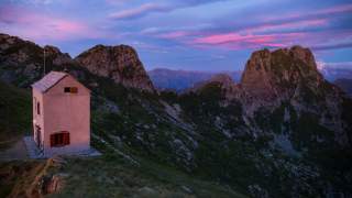 Ein kleines Haus steht auf einem grasbewachsenen Hang inmitten von Berggipfeln bei Sonnenuntergang mit rosa und blauen Wolken am Himmel.