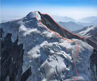 Verschneiter Berggipfel mit Felsformationen und einer roten Linie, die einen Pfad oder eine Route markiert, vor einem klaren blauen Himmel.