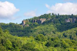 Grüner bewaldeter Hügel mit sichtbaren Felsen unter blauem Himmel mit einigen weißen Wolken.