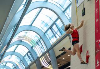 Eine Frau in rotem Top und schwarzen Shorts springt an einer Kletterwand in einer Halle mit großem Glasdach.