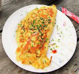 Ein goldbraun gebratener Pfannkuchen, gefaltet und mit fein geschnittenem Schnittlauch bestreut, auf einem weißen Teller mit Blumenmuster, daneben liegt eine Gabel in einer rot-weiß karierten Serviette.