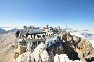 Bergstation mit Aussichtsplattform voller Menschen auf schroffen Felsen in großer Höhe unter klarem Himmel. Im Hintergrund Wolkenmeer und Bergpanorama.