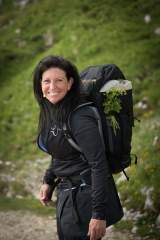 Frau mit schwarzer Outdoor-Bekleidung und großem Wanderrucksack, an dem eine grüne Pflanze befestigt ist, lächelt auf einem Wanderweg vor grün bewachsenem Hügel.