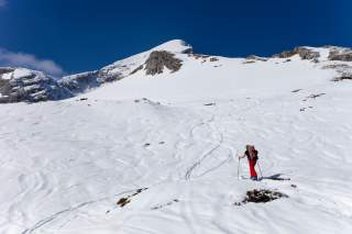 Eine einzelne Person mit roter Skihose und Rucksack steht mit Skiern auf verschneitem Berggelände unter klarem blauem Himmel. Im Hintergrund erhebt sich ein schneebedeckter Gipfel mit sichtbaren Felsen.
