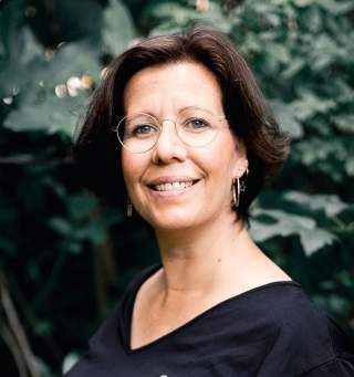 Porträt einer lächelnden Frau mit kurzen braunen Haaren, rundem Brillengestell und schwarzem Oberteil vor unscharfem grünen Hintergrund.