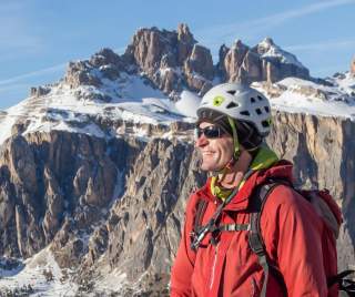 Ein Bergsteiger mit Helm, Sonnenbrille und roter Jacke steht vor schneebedeckten Felsen unter blauem Himmel und lächelt.