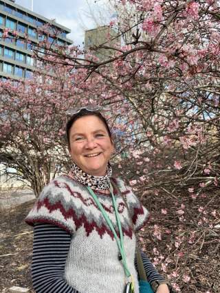 Frau lächelt vor blühendem Baum mit rosa Blüten, im Hintergrund sind Gebäude zu sehen.