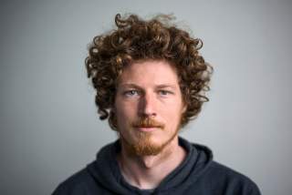 Porträt eines Mannes mit lockigem braunem Haar, rotem Bart und blauem Hoodie vor neutralem Hintergrund.