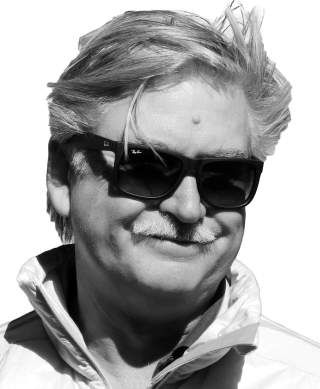Schwarz-weiß-Porträt eines lächelnden Mannes mit Schnurrbart, der eine Sonnenbrille und eine Jacke trägt, vor weißem Hintergrund.