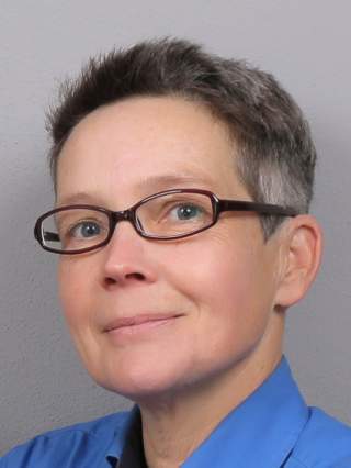 Porträt einer Person mit kurzen braunen Haaren und Brille, die ein blaues Hemd trägt und leicht lächelt.
