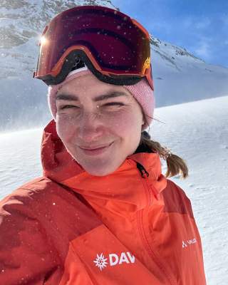 Frau in roter Skijacke und roter Skibrille macht ein Selfie im verschneiten Gebirge bei Sonnenschein.