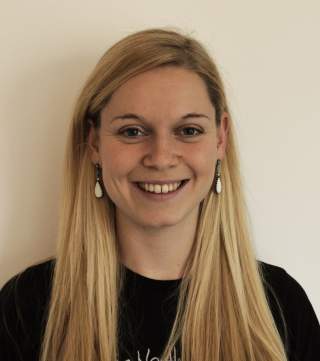 Porträtfoto einer lächelnden Frau mit langen blonden Haaren, die vor einem neutralen hellen Hintergrund steht.
