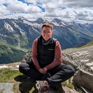 Person mit kurzer Frisur, Brille und Wanderkleidung sitzt lächelnd auf einem Felsen vor einer Berglandschaft mit schneebedeckten Gipfeln und bewölktem Himmel.