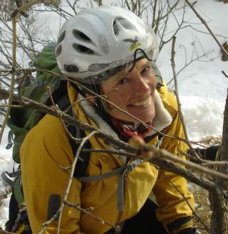 Frau mit Helm, gelber Jacke und Rucksack lächelt in verschneiter Landschaft hinter Ästen hindurch.