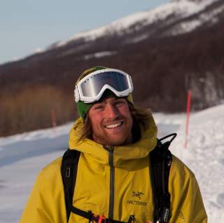 Porträt eines lächelnden Mannes in gelber Winterjacke, Skihelm und Skibrille, stehend auf einer verschneiten Berglandschaft.