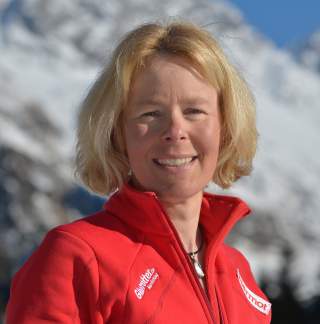 Porträt einer lächelnden Frau mit blonden Haaren, die eine rote Jacke trägt, vor einem Hintergrund aus schneebedeckten Bergen und blauem Himmel.