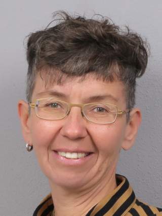 Porträt einer lächelnden Person mit kurzen, gewellten Haaren, die goldene Brille und gestreiftes Hemd trägt, vor grauem Hintergrund.